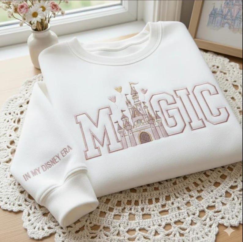 Puede incluir: Sudadera blanca con la palabra "MAGIC" bordada en color marr&oacute;n claro, junto con un gr&aacute;fico de castillo. La manga dice "IN MY DISNEY ERA". La sudadera est&aacute; sobre una superficie blanca de ganchillo.