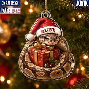 May include: A clear acrylic Christmas tree-shaped ornament featuring a brown and tan patterned snake wearing a red Santa hat with the name RUBY printed on the white brim. The snake is coiled around a red gift box with a gold ribbon and bow.