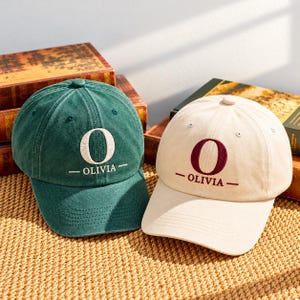 May include: Two baseball caps, one dark green and one cream, each featuring an embroidered large letter O above the name OLIVIA flanked by two horizontal lines. The caps have a structured crown, a curved brim, and a button on the top center.
