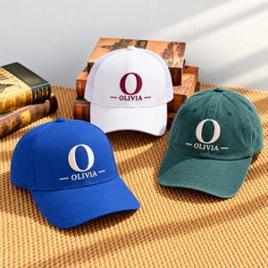 May include: Three baseball caps in blue, white, and dark green. Each cap features a large embroidered letter O above the text OLIVIA in a centered design. The white cap has a mesh back panel, while the blue and green caps are solid fabric.