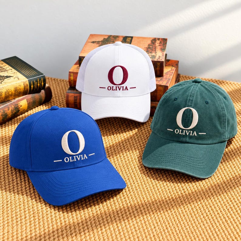 May include: Three baseball caps in blue, white, and dark green. Each cap features a large embroidered letter O above the text OLIVIA in a centered design. The white cap has a mesh back panel, while the blue and green caps are solid fabric.