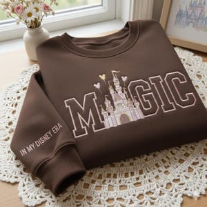 Puede incluir: Una sudadera marr&oacute;n con la palabra "MAGIC" bordada en letras blancas, con un dise&ntilde;o de castillo. La manga dice "IN MY DISNEY ERA". La sudadera est&aacute; doblada sobre un tapete de encaje blanco.