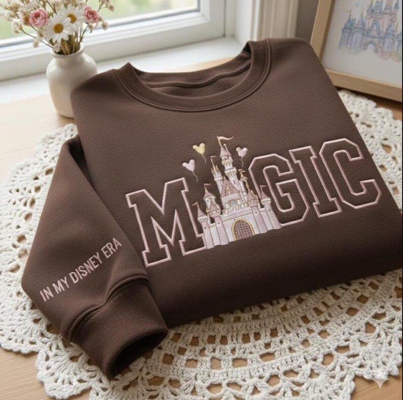 Puede incluir: Una sudadera marr&oacute;n con la palabra "MAGIC" bordada en letras blancas, con un dise&ntilde;o de castillo. La manga dice "IN MY DISNEY ERA". La sudadera est&aacute; doblada sobre un tapete de encaje blanco.