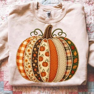 May include: A light beige crewneck sweatshirt featuring a large, central pumpkin graphic. The pumpkin is composed of vertical segments with various patterns, including orange plaid, floral prints, small pumpkins, polka dots, herringbone, and autumn leaves.