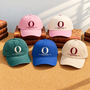 May include: Five baseball caps in pink, cream with a pink brim, dark green, blue, and beige. Each cap features a large embroidered letter O above the name OLIVIA in a serif font, centered on the front panel with horizontal lines on each side.