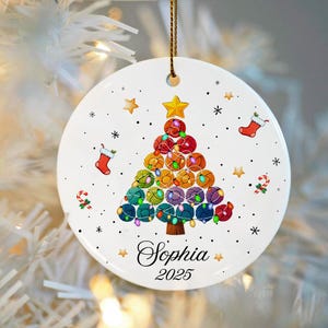 May include: White ceramic Christmas ornament with a colorful yarn ball Christmas tree design. The ornament includes the name "Sophia" and the year "2025". Additional details include stars, snowflakes, candy canes, and stockings.