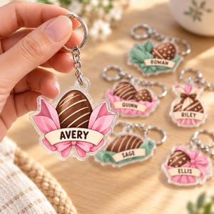 May include: A clear acrylic keychain featuring a brown chocolate egg graphic with white stripes, wrapped in pink decorative paper with a banner displaying the name AVERY. Other similar keychains show names ROWAN, QUINN, RILEY, SAGE, and ELLIS.