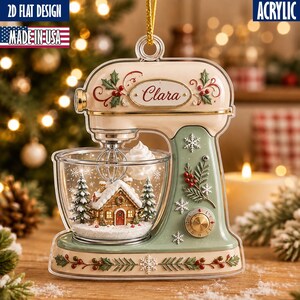 May include: An acrylic stand mixer ornament featuring a cream and sage green finish with holly, snowflake, and berry accents. The mixing bowl contains a miniature gingerbread house, pine trees, and snow. The name Clara is printed on the front.