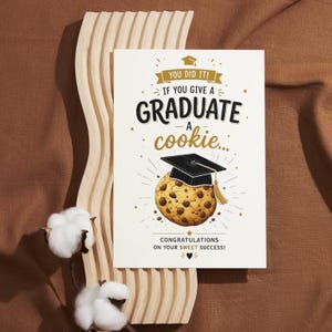 May include: A graduation greeting card with the text "YOU DID IT! IF YOU GIVE A GRADUATE A cookie..." and "CONGRATULATIONS ON YOUR SWEET SUCCESS!". The card features a cookie wearing a graduation cap. The card is on a wooden stand.