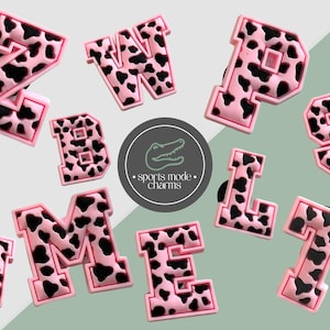 May include: A collection of pink and black cow print alphabet charms. The letters are in a bold, block style with a raised border. The image also includes a circular logo with the text "sports mode charms".