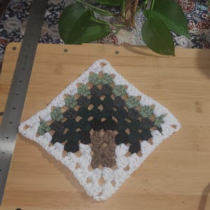 May include: A crocheted square with a green, brown, and white tree design. The square is made with white yarn and has a brown trunk and green leaves.