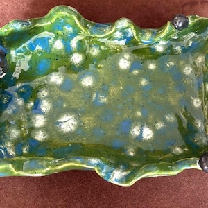 Puede incluir: Un plato de cerámica verde con un esmalte moteado azul y blanco. El plato tiene un borde ondulado y una pequeña figura de perro en un lado.