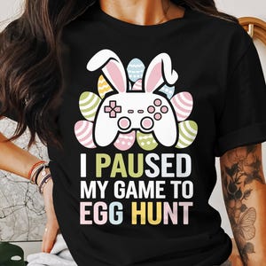 May include: Black t-shirt with a white video game controller and bunny ears surrounded by colorful Easter eggs. The text "I paused my game to egg hunt" is printed below the controller.