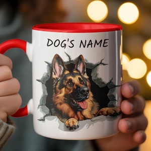 May include: White ceramic mug with a red rim, featuring a German Shepherd dog peeking out of a broken wall. The mug has the text "DOG'S NAME" printed on the front.