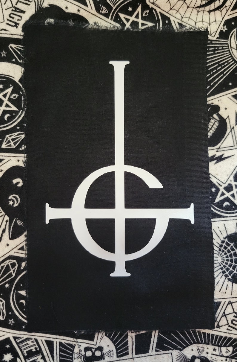 K&ouml;nnte beinhalten: Schwarzer Stoff-Patch mit einem wei&szlig;en umgekehrten Kreuz, das von einem Kreis umgeben ist. Das Symbol wird oft mit der Band Ghost in Verbindung gebracht.