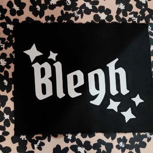 May include: Black rectangle with the word "Bleh" in white, stylized gothic font, and white star accents. The background is a floral pattern in black and tan.