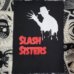 May include: Black and red iron-on patch with a white silhouette of a horror movie character with a knife and the text "SLASH SISTERS".