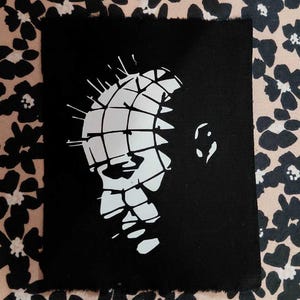 Peut inclure: Impression artistique rectangulaire noire avec un graphisme blanc d'un visage quadrillé et d'épingles. L'impression a un bord brut. Le fond est un motif floral noir et beige.
