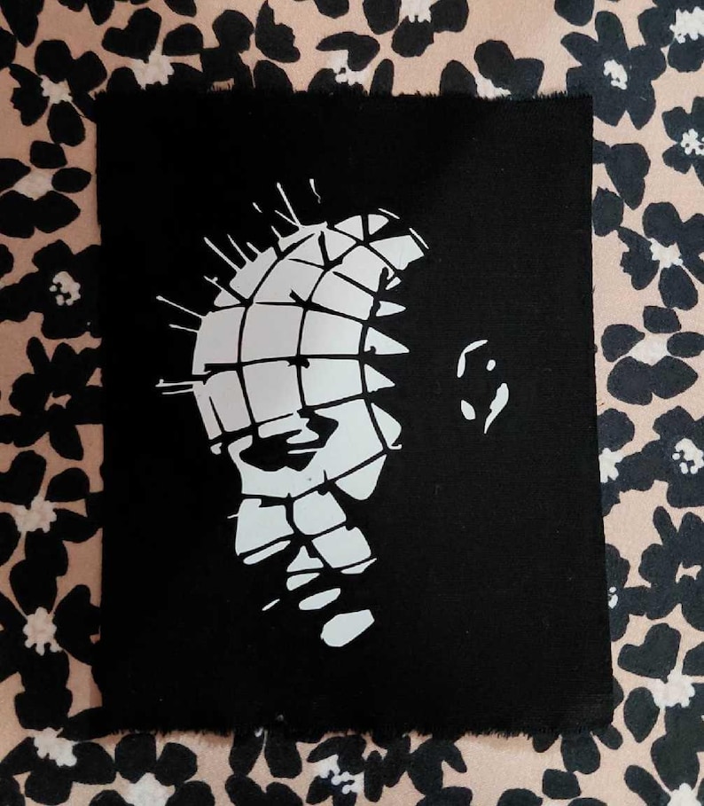 May include: Black rectangular art print with a white graphic of a face with a grid pattern and pins. The print has a rough edge. The background is a floral pattern in black and tan.