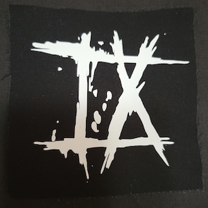Op de afbeelding: Witte patch met een gestileerd "IX" ontwerp op een zwarte achtergrond. De patch is gemaakt van stof en heeft een ruw, getextureerd oppervlak.