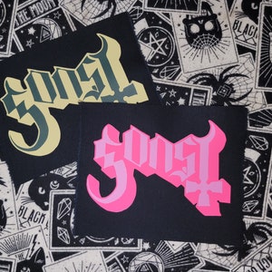May include: Two black fabric patches with the word "Ghost" in a gothic font. One patch has the word in a light green color, and the other in pink. The patches are on a black and white patterned background.