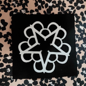 May include: A black square patch featuring a white, stylized star design with a floral border. The patch is set against a patterned background of black floral shapes on a tan surface.