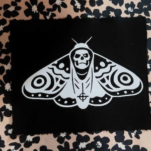 May include: A black patch featuring a white moth design with a skull face and a cross symbol. The moth has detailed wing patterns. The patch is set against a patterned background.