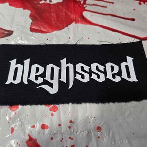 May include: A black rectangular patch with the word "bleghsssed" in white gothic font. The patch is on a white surface with red splatters.