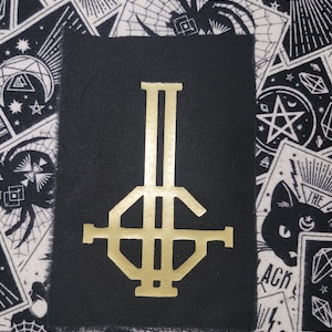 Pode incluir: Um emblema retangular de tecido preto com um logótipo serigrafado em dourado metálico. O design consiste numa linha vertical cruzada por uma barra horizontal e uma forma octogonal central, formando um emblema estilizado no tecido.