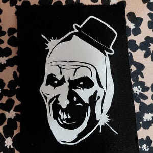 May include: A black patch featuring a white graphic of a sinister clown face wearing a small hat. The clown has a wide, toothy grin and piercing eyes. The patch has a raw edge and is set against a floral background.