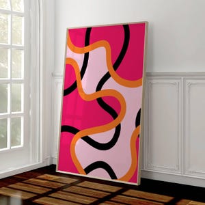 May include: A framed abstract art print featuring a vibrant design. The artwork showcases flowing lines in black, orange, and pink against a light pink background. The frame is a light wood color, adding a natural touch to the modern design.