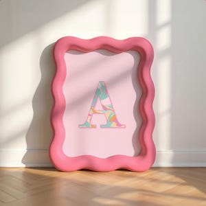Puede incluir: Impresión enmarcada en rosa con la letra "A". El marco tiene un borde ondulado. La letra "A" presenta un patrón abstracto colorido en colores pastel. El fondo es de color rosa claro.