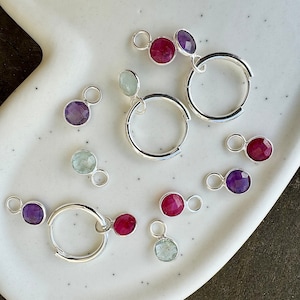 May include: A set of silver hoop earrings with gemstone charms. The charms are in various colors, including purple, red, and green. The earrings are displayed on a white plate.