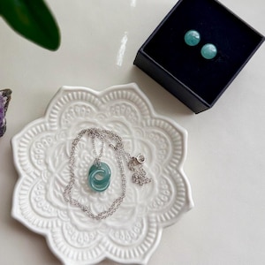 May include: A silver necklace with a light blue pendant rests on a white decorative plate. Matching light blue stud earrings are in a black box. The jewelry is displayed on a white surface, with a green plant in the background.
