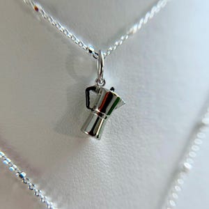 May include: A silver-colored necklace featuring a small charm shaped like a stovetop espresso maker. The charm has a handle and spout, and hangs from a delicate chain. The background is a soft, neutral color.