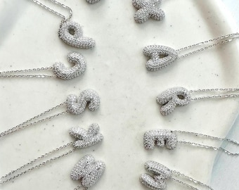 May include: A set of 12 silver necklaces with letter pendants. Each pendant is made of cubic zirconia stones and is shaped like a puffy letter. The letters are A, B, C, D, E, F, G, H, I, J, K, and L.