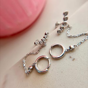 May include: A pair of silver-toned earrings featuring a small hoop connected by a delicate chain to a curved bar stud. The curved bar is set with four small, clear, round-cut stones, and a single clear stone is embedded on the hoop attachment point.