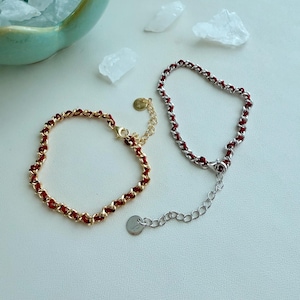 May include: Two chain bracelets featuring red cord woven through metal links. One bracelet is gold-toned with a circular charm, and the other is silver-toned with a circular charm. Both include an adjustable extender chain with a lobster clasp.