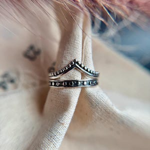May include: A silver-toned ring with a crown design. The ring features two bands, one with small, evenly spaced dots and the other with a series of small, dark circles. The crown portion has a series of small dots. The ring is set against a neutral fabric background.