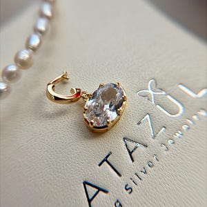 May include: A gold-toned hoop earring featuring a dangling oval-cut clear gemstone in a prong setting. The jewelry is positioned on a textured surface with the text LATAZUL sterling silver jewelry printed in dark blue letters nearby.