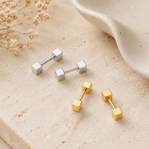 May include: A collection of barbell-shaped stud earrings. Two pairs are silver-toned, and two pairs are gold-toned. Each earring features a small, square cube at each end of a straight bar. The earrings are displayed on a light-colored, textured surface.