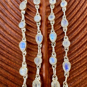May include: Three silver chains with teardrop-shaped moonstone beads. The chains are on a brown wooden background.