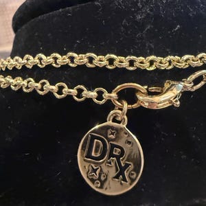 Pode incluir: Colar dourado com um pingente redondo. O pingente apresenta as letras "DRX" e detalhes em forma de estrela. O colar &eacute; exibido em uma superf&iacute;cie preta.