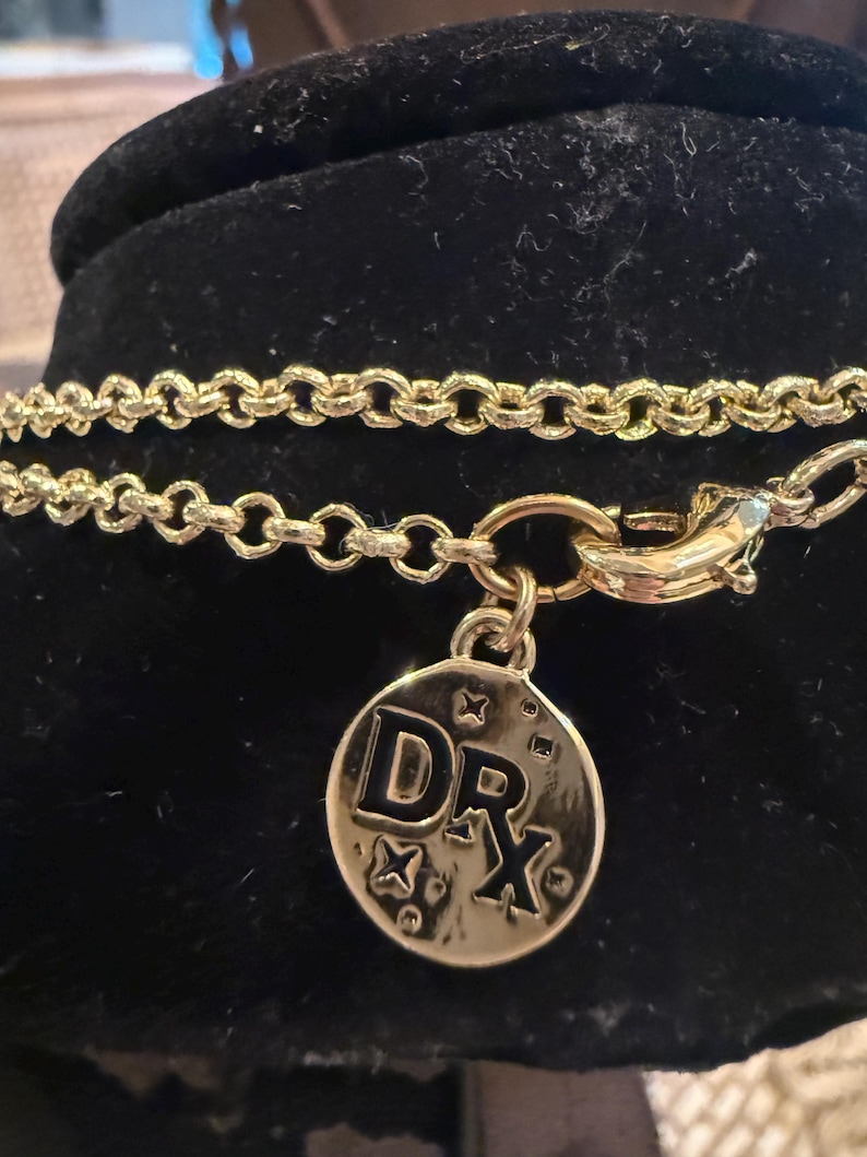 Pode incluir: Colar dourado com um pingente redondo. O pingente apresenta as letras "DRX" e detalhes em forma de estrela. O colar &eacute; exibido em uma superf&iacute;cie preta.