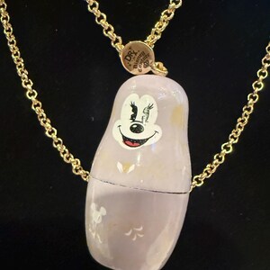 Pode incluir: Colar com uma boneca russa pintada &agrave; m&atilde;o, de cor lil&aacute;s claro, com o rosto sorridente da Minnie Mouse. A boneca est&aacute; suspensa por uma corrente dourada. O colar tem um fecho com o texto "DR. Disney Couture".