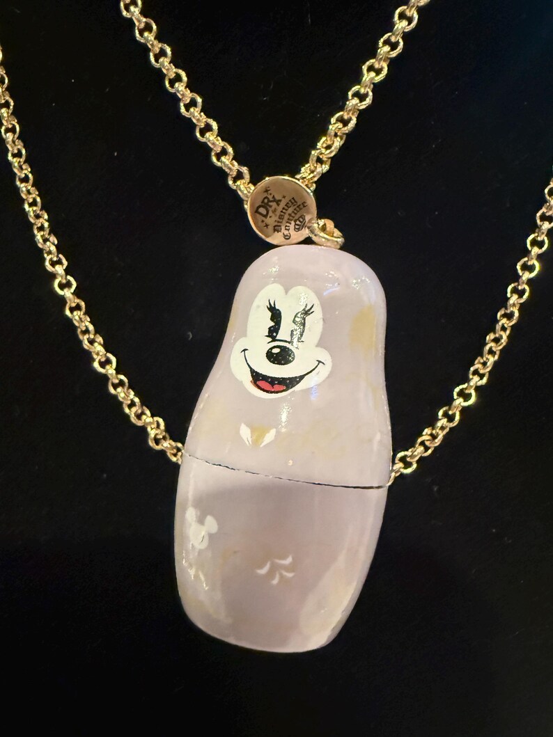 Pode incluir: Colar com uma boneca russa pintada &agrave; m&atilde;o, de cor lil&aacute;s claro, com o rosto sorridente da Minnie Mouse. A boneca est&aacute; suspensa por uma corrente dourada. O colar tem um fecho com o texto "DR. Disney Couture".
