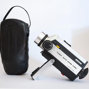 Könnte beinhalten: Vintage Canon Auto Zoom 318M Videokamera mit schwarzer Ledertragetasche. Die Kamera ist silber und schwarz mit Zoomobjektiv und Griff. Die Tasche ist schwarz mit einem Griff.
