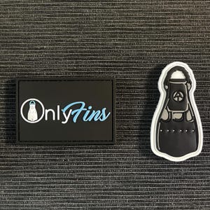 Pode incluir: Dois patches pretos com texto branco e azul e um patch em forma de barbatana preto e branco. O patch retangular tem o texto "Only Fins" com um gráfico de farol. O patch em forma de barbatana tem um design de barbatana de mergulho.