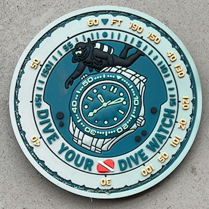 Puede incluir: Un parche circular en color verde azulado y verde claro con un gráfico central de un reloj de pulsera y un buceador. El texto DIVE YOUR DIVE WATCH está impreso en la parte inferior. El borde exterior muestra marcas de profundidad en pies, incluyendo 30, 40, 50, 60, 150 y 190.