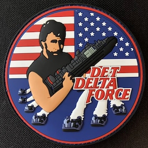 Peut inclure: Un patch circulaire bleu, rouge et blanc avec une image de bande dessinée d'un homme tenant une grosse arme. L'homme porte un débardeur noir et a une barbe. Le texte "DET DELTA FORCE" est imprimé en lettres rouges et blanches sur le patch.